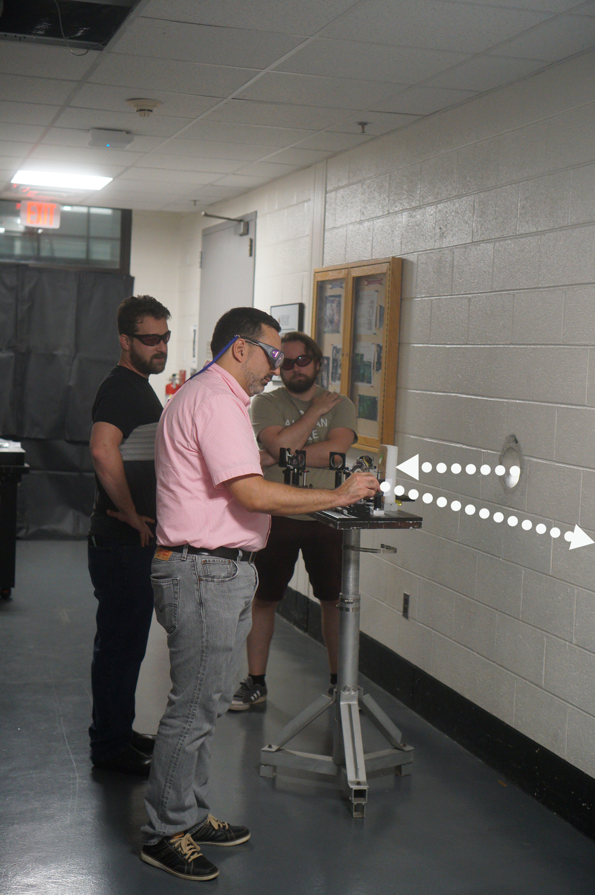  Left to right Eric Rosenthal, a physicist at the U.S. Naval Research Laboratory; Anthony Valenzuela, a physicist at the U.S. Army Research Lab; and Goffin align optics at a porthole in the wall in order to send the laser beam from the lab down the hallway. The white dotted lines show the approximate beam path before and after the optics redirected it. 